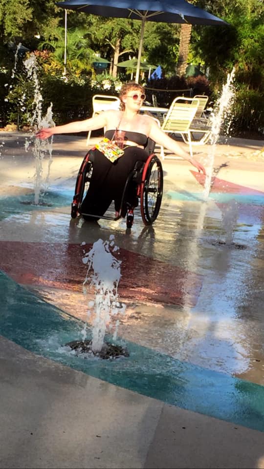 Samm sitting in her wheelchair with arms spread out in a splash pad fountain.